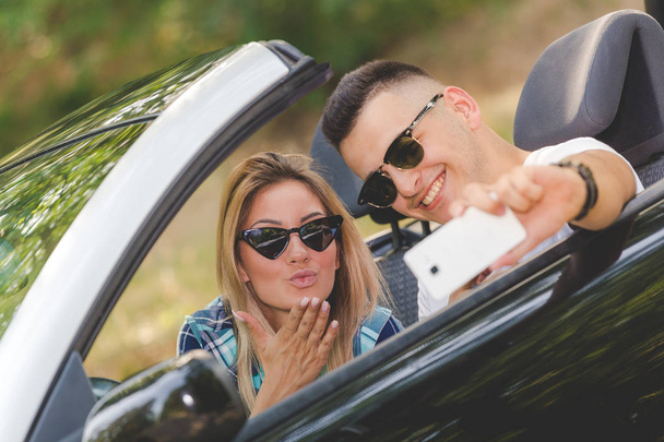 Young couple taking selfie with smartphone while sitting in convertible car. Travel and technology concepts. - Photo, Image