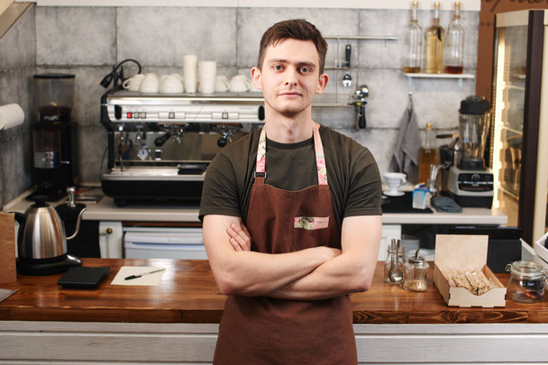 man barista with crossed arms leaning on wooden bar surface at workplace in coffee shop - Foto, Imagem