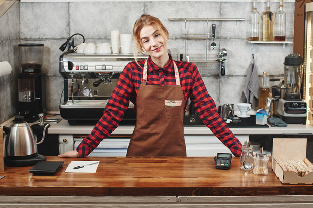 smiling attractive barista standing and looking at camera at workplace in coffee shop - Фото, зображення