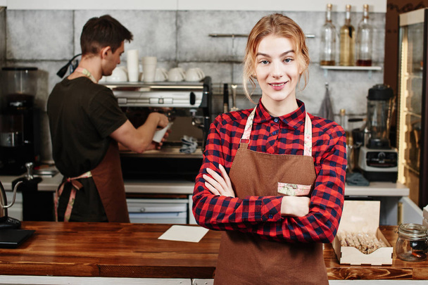 female barista with crossed arms looking at camera while standing in front of bar with colleague near coffee machine on background  - Фото, зображення