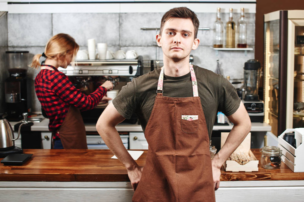 handsome barista looking at camera and standing in front of bar with colleague near coffee machine on background  - Photo, Image