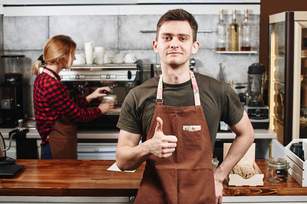 handsome barista looking at camera and standing in front of bar with colleague near coffee machine on background  - Photo, image