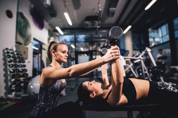 νεαρή γυναίκα, άσκηση με βάρη, ενώ της γυναικείας προσωπικό γυμναστή βοηθά στο γυμναστήριο - Φωτογραφία, εικόνα