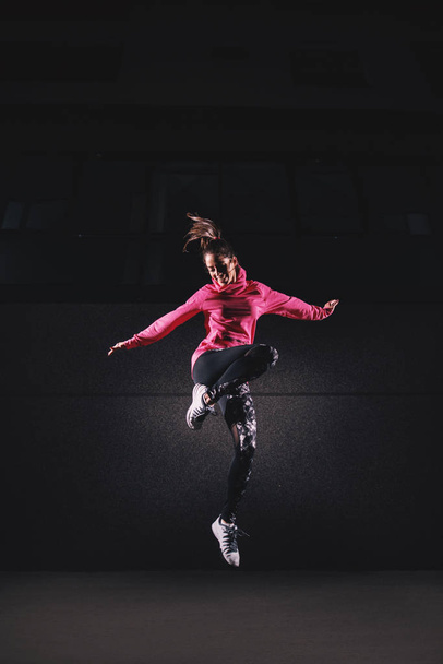 portrait of young slim woman in sportswear jumping against dark wall - Foto, afbeelding