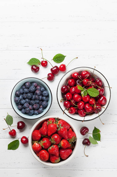 Fresh cherries, blueberries and strawberries in bowls on white wooden background. Top view with space for your text - Zdjęcie, obraz