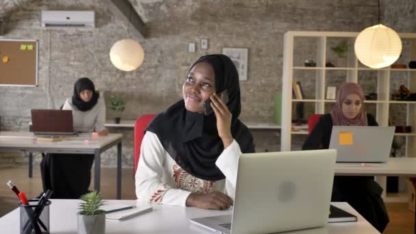 Young black muslim women in hijab talking on phone, smiling and laughing in office, three pretty muslim womens working on laptop - Video