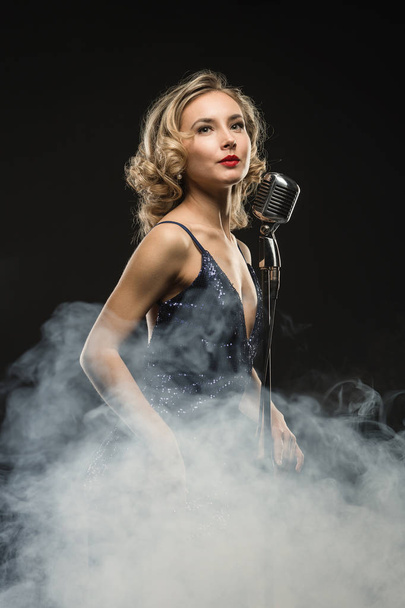 Sexy young girl singer singing with silver retro microphone on black background with stage smoke - 写真・画像