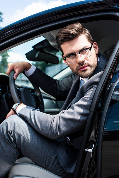pensive businessman in eyeglasses looking away while sitting in car - Foto, Imagen