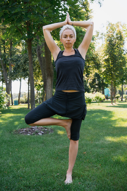 Frau praktiziert Yoga in Baum-Pose (vrksasana) und blickt auf Gras im Park in die Kamera - Foto, Bild