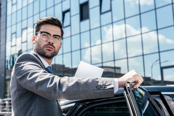 side view of confident businessman with papers in hands standing near car on street - Photo, Image