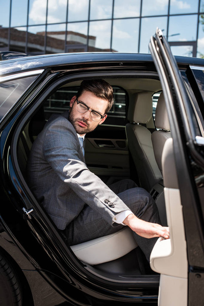 Zijaanzicht van peinzende zakenman op zoek weg zittend op de achterbank met geopende auto deur - Foto, afbeelding