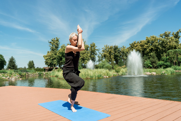 спортсменка практикующая йогу и стоящая на одной ноге на коврике для йоги возле реки в парке
 - Фото, изображение