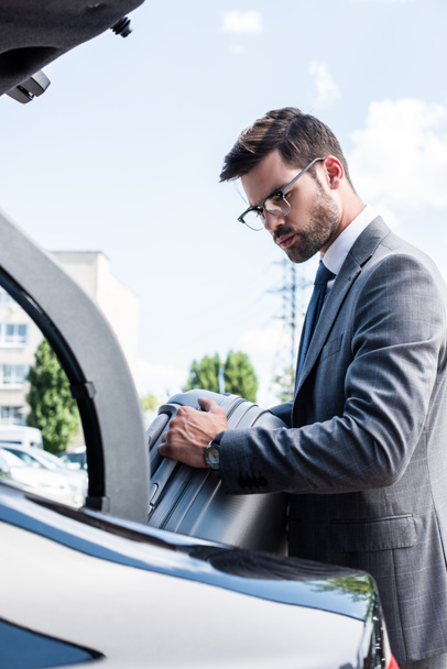 handsome businessman in eyeglasses putting wheeled bag in car trunk at street  - Fotó, kép