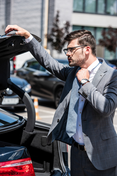 young businessman in eyeglasses closing car trunk at street - 写真・画像