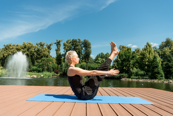 вид збоку на спортсменку, що практикує йогу на йозі-маті біля річки в парку
 - Фото, зображення