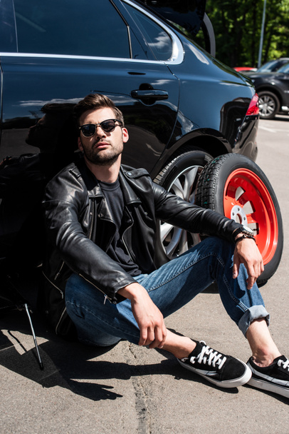 tired stylish man in sunglasses sitting near broken car at street  - Photo, Image