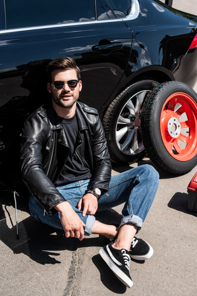 smiling stylish man in sunglasses sitting near broken car at street - Foto, immagini