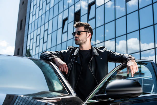 młody człowiek stylowy w skórzanej kurtce i okulary przeciwsłoneczne, patrząc od obiektu w pobliżu samochodów ulicy - Zdjęcie, obraz