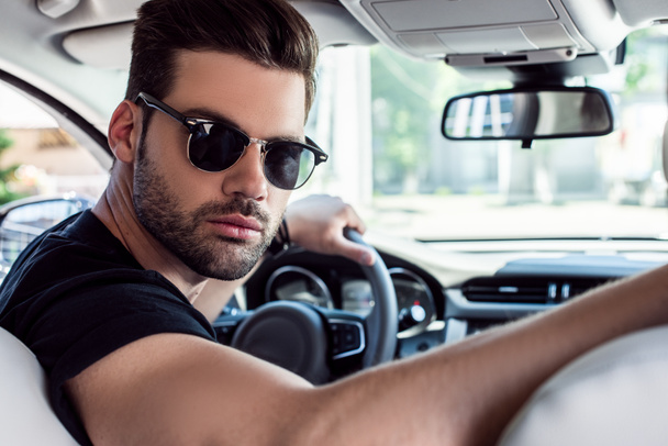 portrait of serious stylish man in sunglasses looking back while driving car  - Photo, Image