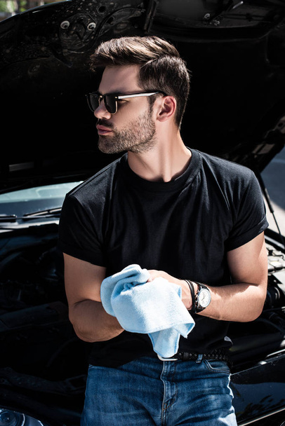 young man in sunglasses wiping hands by rag near car with opened bonnet  - Foto, imagen