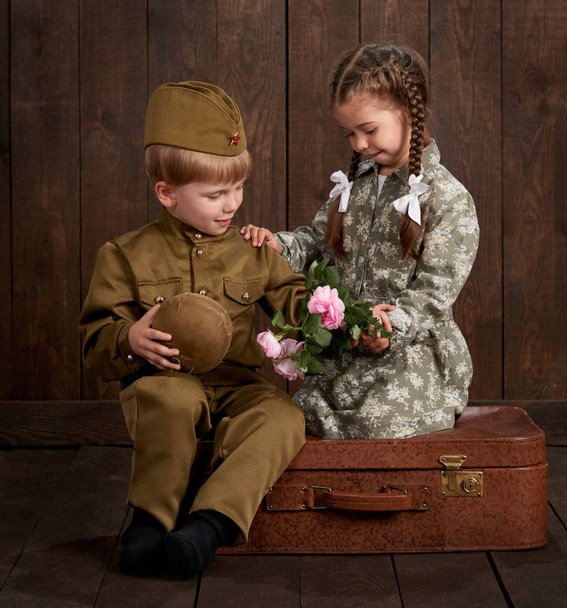 Çocuk Çocuk retro askeri üniformalı asker ve kız pembe elbiseli oturma eski bavul, koyu ahşap arka plan, retro tarzı olarak giymiş - Fotoğraf, Görsel