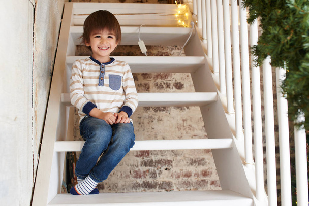 kleine jongen zit op houten witte trap in appartement ingericht voor Kerstmis - Foto, afbeelding