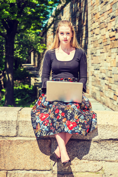 Jovem estudante universitário adolescente americano estudando em Nova York, vestindo t-shirt manga comprida preta, skit modelado, sapatos de salto alto, sentado à beira da parede de rocha no campus sob o sol, trabalhando no computador portátil
 - Foto, Imagem