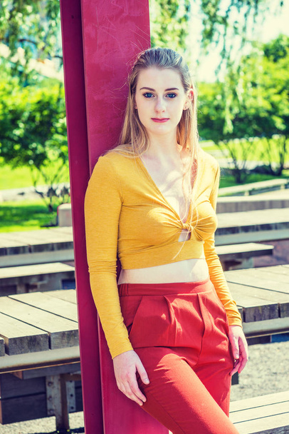 Teenage Girl Spring Fashion in New York. Young American woman with long blonde hair, white skin, wearing yellow long sleeve deep v neck fit crop top, red orange pants, standing by red pole at park - Foto, imagen