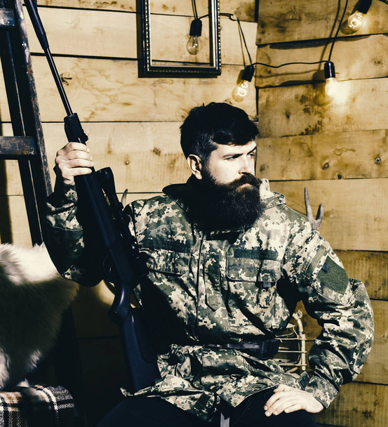 Man with beard wears camouflage clothing in wooden interior background. Hunter, brutal hipster with gun in his hand ready for hunting. Gamekeeper concept. Macho on strict face at gamekeepers house. - Φωτογραφία, εικόνα