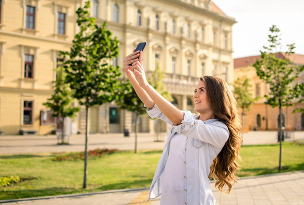 Beautiful young brunette with long hair taking smiling selfi in the town - Fotó, kép