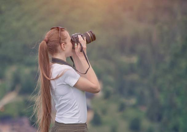 Girl with camera stands on a hill and photographing nature. Forest on background - Photo, Image