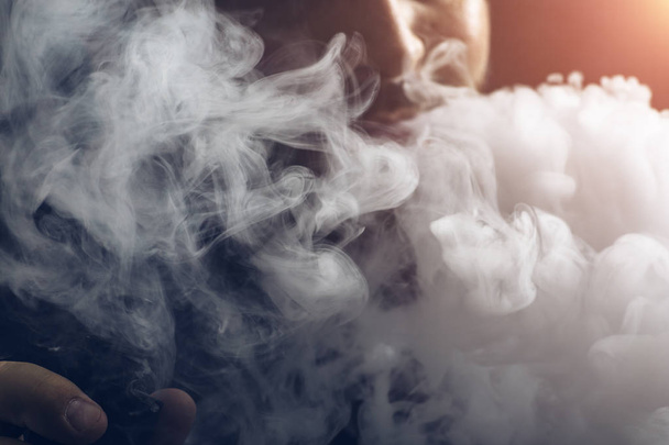 Homem vape e-cigarro com e-líquido, close-up, respira grande nuvem de vapor ou vapor. Conceito de vaporização, efeito de luz
 - Foto, Imagem