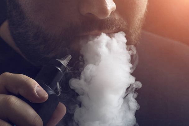 Man vape e-cigarette with e-liquid, close-up, breathes out large cloud of steam or vapor. Vaping concept, light effect - Foto, afbeelding