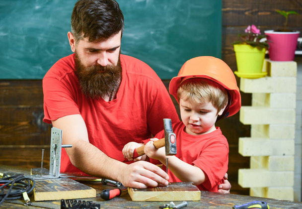 Handyman concept. Father with beard teaching little son to use tools, hammering, chalkboard on background. Boy, child busy in protective helmet learning to use hammer with dad - Φωτογραφία, εικόνα