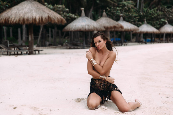 A gentle, attractive young girl with a slender figure and a tanned body sitting on white sand on her knees, gently embraces herself and covers her bare chest with her hands. - Foto, afbeelding