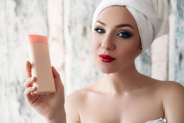 Primer plano de una hermosa joven que acaba de salir de la ducha, mostrando su maquillaje, Mujer con una toalla en el pelo, hermoso maquillaje, labios rojos, sostiene una botella de cosméticos en la mano sin
 - Foto, Imagen