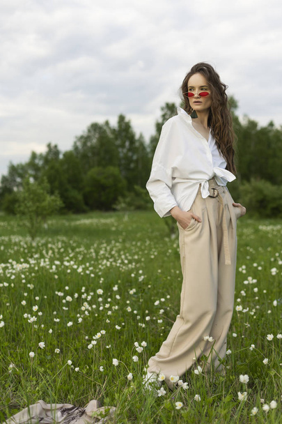 Beautiful stylish young girl wearing a trendy white shirt, beige bloomers and red sunglasses poses outdoors on the meadow full of white wild flowers. Commercial and advertisement design. Copy space - Photo, image