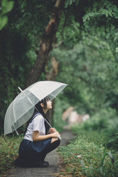 Porträt eines asiatischen Schulmädchens, das bei Regen mit Regenschirm auf dem Naturweg sitzt - Foto, Bild