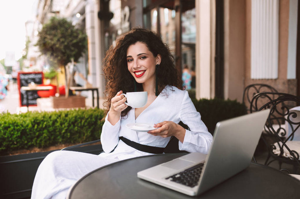 Mooie lachende dame met donkere krullend haar in wit kostuum zittend aan tafel met laptop en kopje koffie in de handen terwijl vrolijk in de camera kijken in café op straat - Foto, afbeelding