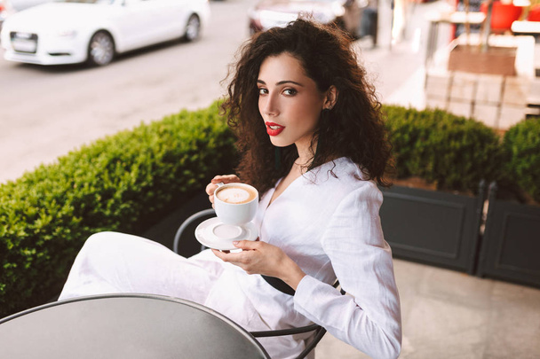 Beautiful lady with dark curly hair in white costume sitting at the table with cup of coffee in hands and thoughtfully looking in camera in cafe on street - Photo, image