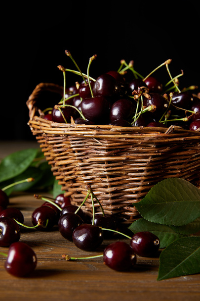 ripe harvested cherries in rustic basket with leaves on wooden table and on black - Photo, Image