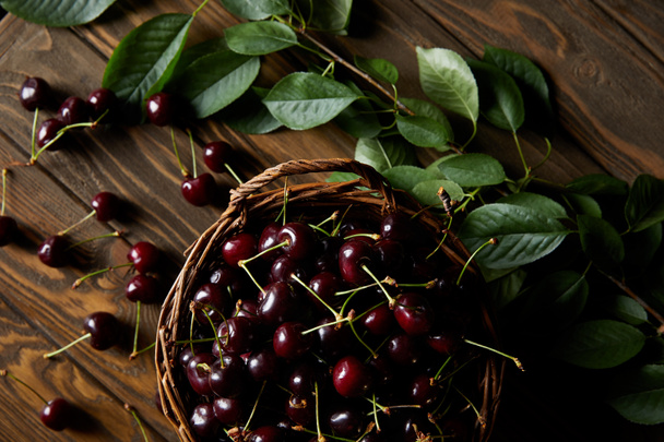 top view of fresh red sweet cherries in rustic basket on wooden table with leaves - Fotó, kép