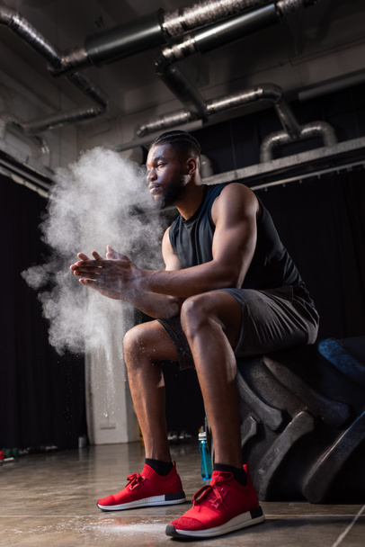 low angle view of african american sportsman sitting in tyre and applying talcum powder on hands - Foto, Imagen