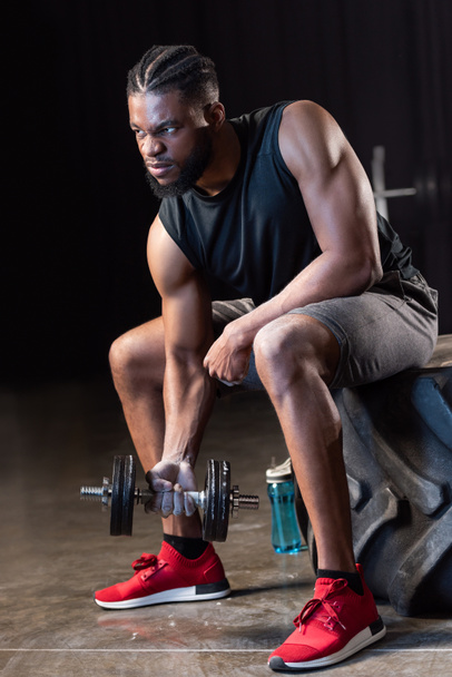 muscular african american sportsman sitting on tyre and exercising with dumbbell  - Zdjęcie, obraz