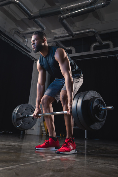 low angle view of muscular african american sportsman lifting barbell and looking away in gym  - 写真・画像