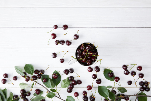 top view of fresh ripe cherries in cup on white wooden tabletop with leaves branch - Fotografie, Obrázek