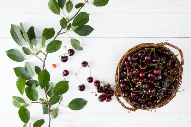top view of fresh ripe cherries in rustic basket on white wooden tabletop with leaves branch - Fotografie, Obrázek