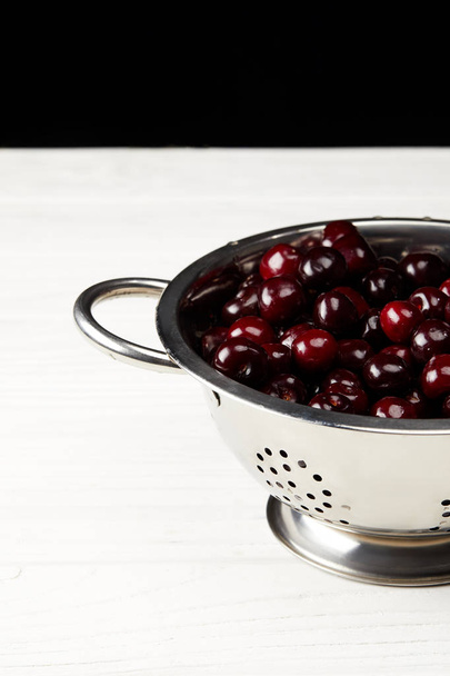 red ripe sweet cherries in metal colander on white wooden surface and on black - Foto, imagen
