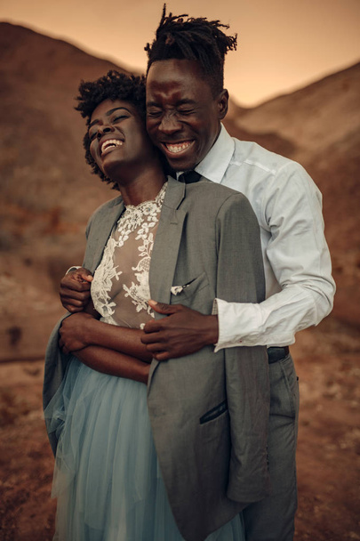 Μαύρο γαμπρός αγκαλιάζει τη νύφη του ντυμένος με σακάκι του στο φαράγγι ενάντια όμορφο τοπίο κατά το ηλιοβασίλεμα. Γελάνε χαρούμενα. Κινηματογράφηση σε πρώτο πλάνο. - Φωτογραφία, εικόνα