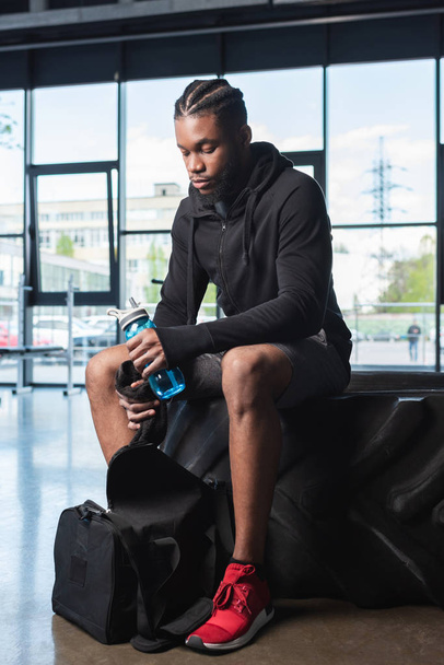 young african american sportsman holding bottle of water and towel while sitting in tyre in gym - Foto, Imagen